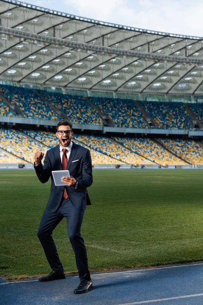 Jovem empresário de terno e óculos usando tablet digital e mostrando gesto yeas no estádio, conceito de apostas esportivas — Fotografia de Stock