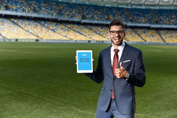 KYIV, UCRANIA - 20 DE JUNIO DE 2019: joven empresario feliz con traje y gafas sosteniendo tableta digital con aplicación skype y mostrando el pulgar hacia arriba en el estadio - foto de stock