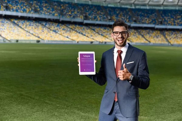 KYIV, UKRAINE - JUNHO 20, 2019: jovem empresário feliz de terno e óculos segurando tablet digital com aplicativo instagram e mostrando o polegar no estádio — Fotografia de Stock