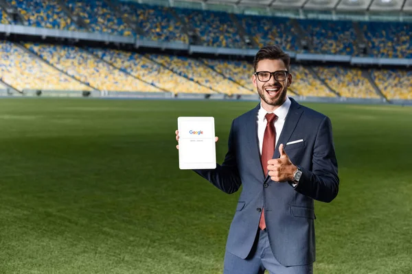 KYIV, UKRAINE - JUNE 20, 2019: happy young businessman in suit and glasses holding digital tablet with google app and showing thumb up at stadium — Stock Photo