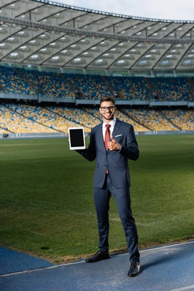 Visão comprimento total do jovem empresário feliz em terno e óculos segurando tablet digital com tela em branco e mostrando o polegar para cima no estádio — Fotografia de Stock