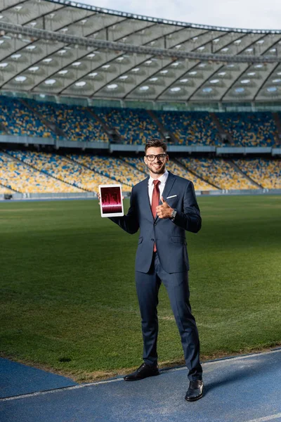 Full length view of happy young businessman in suit and glasses holding digital tablet with trading app and showing thumb up at stadium — Stock Photo