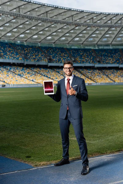 Vista completa de feliz joven hombre de negocios en traje y gafas que sostienen la tableta digital con aplicación de comercio y mostrando el pulgar hacia arriba en el estadio — Stock Photo