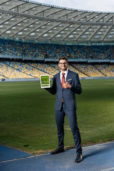 Full length view of happy young businessman in suit and glasses holding digital tablet with healthcare app and showing thumb up at stadium — Stock Photo