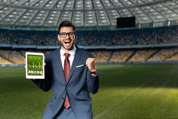 Jovem empresário alegre em terno e óculos segurando tablet digital com aplicativo de saúde e mostrando sim gesto no estádio — Fotografia de Stock