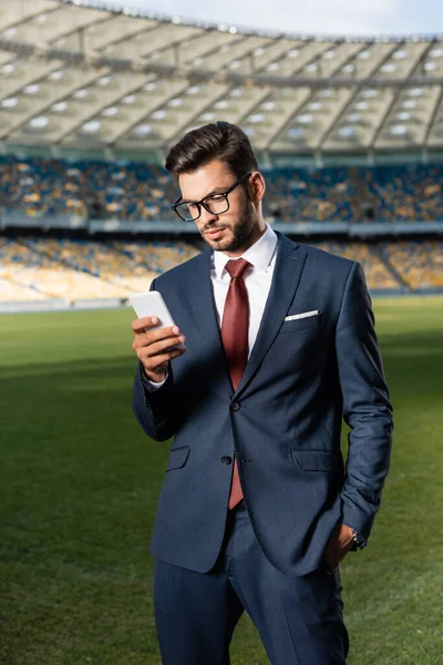 Young businessman in suit and glasses using smartphone at stadium — Stock Photo