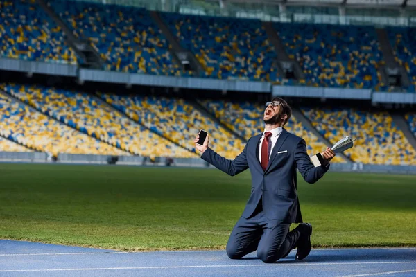 Jeune homme d'affaires en costume debout sur les genoux avec smartphone et trophée au stade, concept de paris sportifs — Photo de stock