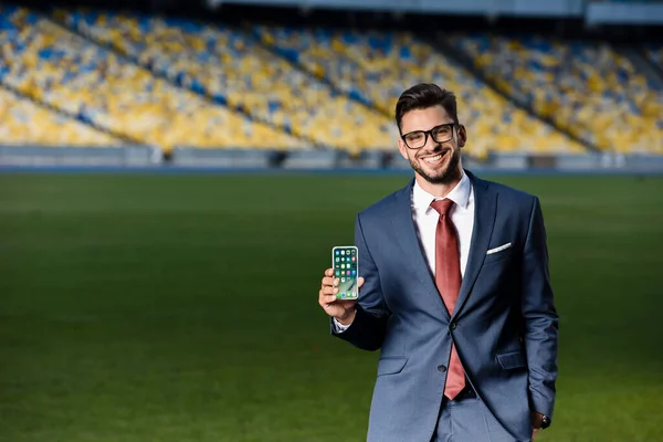 KYIV, UCRANIA - 20 de junio de 2019: joven hombre de negocios sonriente con traje y gafas que sostiene el teléfono inteligente con aplicaciones en la pantalla en el estadio - foto de stock