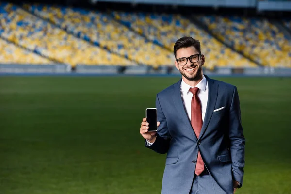 Jovem empresário sorridente de terno e óculos segurando smartphone com tela em branco no estádio — Fotografia de Stock