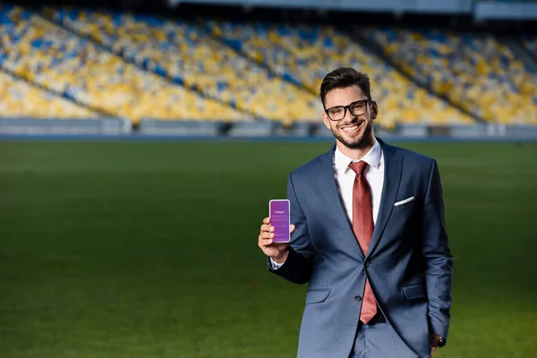 KYIV, UCRANIA - 20 de junio de 2019: joven hombre de negocios sonriente con traje y gafas que sostiene el teléfono inteligente con la aplicación instagram en el estadio - foto de stock