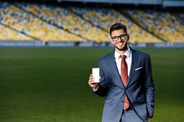 Smiling young businessman in suit and glasses holding smartphone with blank screen at stadium — Stock Photo