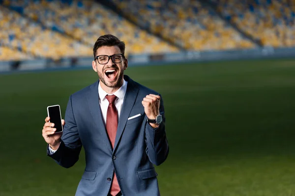 Excité jeune homme d'affaires en costume et lunettes tenant smartphone avec écran blanc et montrant geste oui au stade — Photo de stock