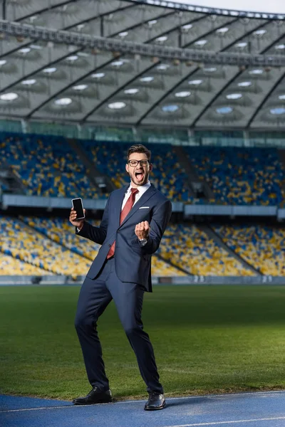 Полный вид возбужденного молодого бизнесмена в костюме и очках, держащего смартфон с пустым экраном и показывающего жест 