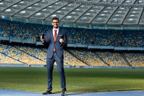 Smiling young businessman in suit and glasses holding smartphone with blank screen and showing yes gesture at stadium — Stock Photo