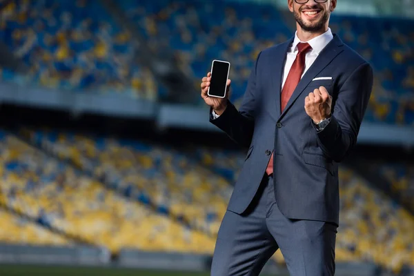 Обрезанный вид улыбающегося молодого бизнесмена в костюме, держащего смартфон с пустым экраном и показывающего жест 
