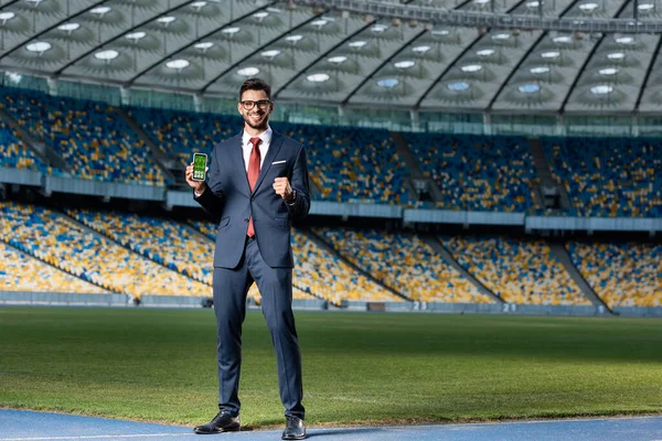 Smiling young businessman in suit and glasses holding smartphone with healthcare app and showing yes gesture at stadium — Stock Photo