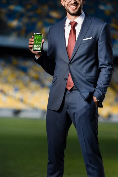 Обрезанный вид молодого бизнесмена в костюме с рукой в кармане, держащего смартфон с приложением для здравоохранения на стадионе — стоковое фото