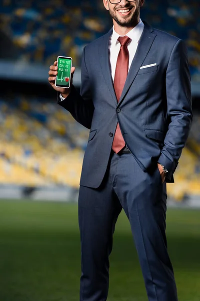 Cropped view of young businessman in suit with hand in pocket holding smartphone with cardiological app at stadium — Stock Photo