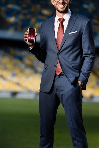 Cropped view of young businessman in suit with hand in pocket holding smartphone with trading app at stadium — Stock Photo