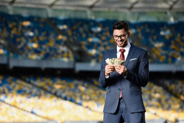 Smiling young businessman in suit and glasses with money at stadium — Stock Photo