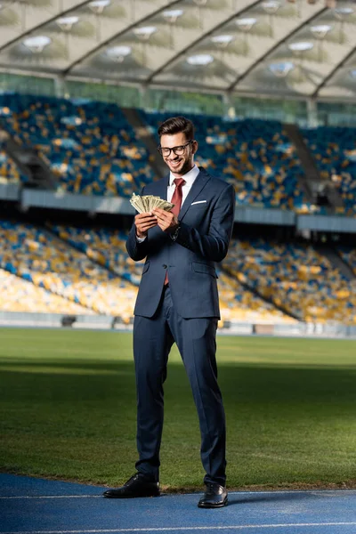 Улыбающийся молодой бизнесмен в костюме и очках с деньгами на стадионе — стоковое фото