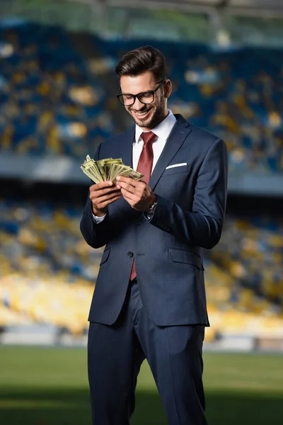 Усміхнений молодий бізнесмен в костюмі і окулярах з грошима на стадіоні — стокове фото