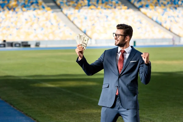Smiling young businessman in suit and glasses with money showing yeas gesture at stadium — Stock Photo