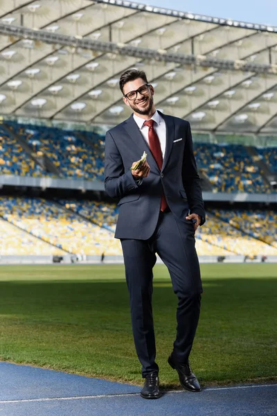 Sorridente giovane uomo d'affari in giacca e cravatta e occhiali dando soldi allo stadio — Foto stock