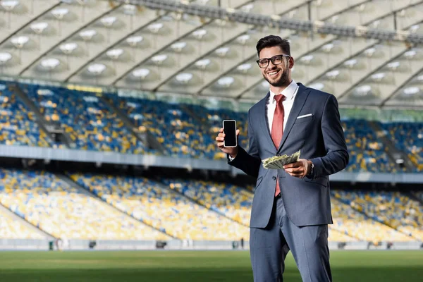 Sonriente joven hombre de negocios en traje y gafas con dinero mostrando teléfono inteligente con pantalla en blanco en el estadio - foto de stock