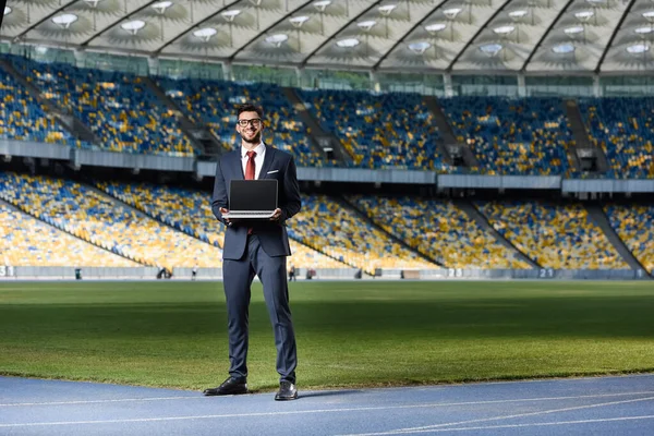 Smiling young businessman in suit with laptop at stadium — Stock Photo