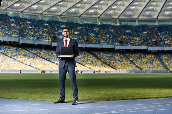 Sonriente joven hombre de negocios en traje con portátil en el estadio - foto de stock