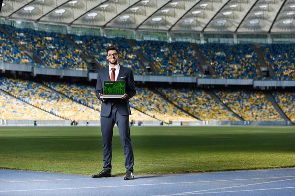Sonriente joven hombre de negocios en traje mostrando portátil con sitio web de comercio en línea en el estadio - foto de stock