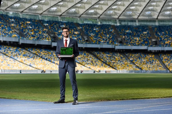 Smiling young businessman in suit showing laptop with online trade website at stadium — Stock Photo