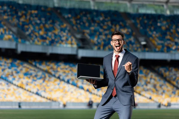 Счастливый молодой бизнесмен в костюме держит ноутбук с чистым экраном и показывает жест 
