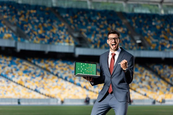 Heureux jeune homme d'affaires en costume tenant ordinateur portable avec terrain de football et formation à l'écran et montrant geste oui au stade — Photo de stock