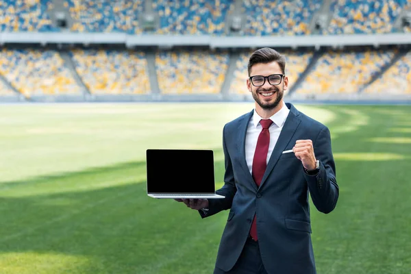 Lächelnder junger Geschäftsmann im Anzug zeigt Ja-Geste, während er Laptop mit leerem Bildschirm im Stadion hält — Stockfoto