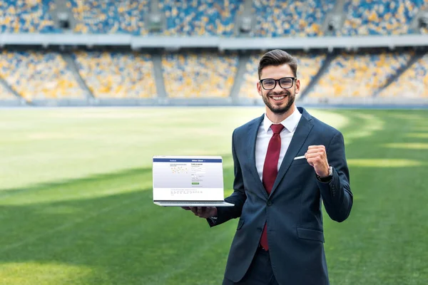 KYIV, UKRAINE - JUNE 20, 2019: smiling young businessman in suit showing yes gesture while holding laptop with facebook website at stadium — Stock Photo