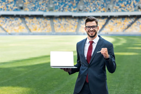 Sonriente joven hombre de negocios en traje mostrando sí gesto mientras sostiene el ordenador portátil con pantalla en blanco en el estadio - foto de stock