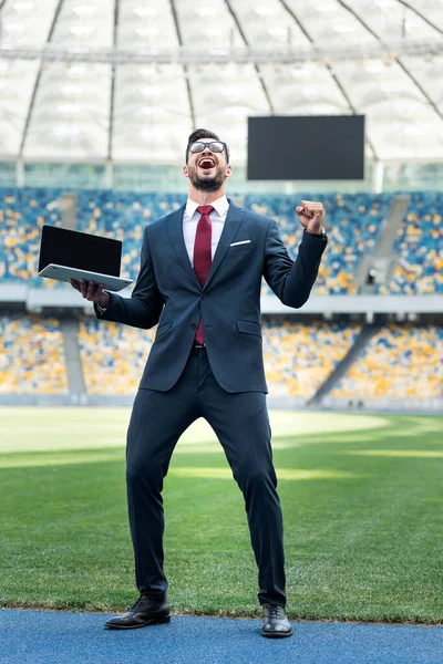 Feliz joven hombre de negocios en traje mostrando sí gesto y la celebración de la computadora portátil con pantalla en blanco en el estadio - foto de stock