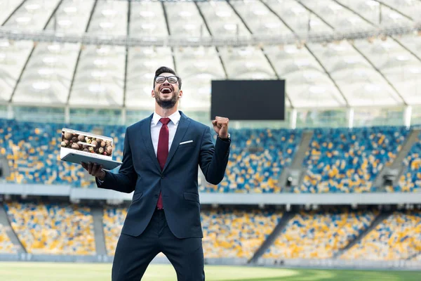 KYIV, UKRAINE - JUNE 20, 2019: low angle view of happy young businessman in suit showing yes gesture and holding laptop with depositphotos website at stadium — Stock Photo