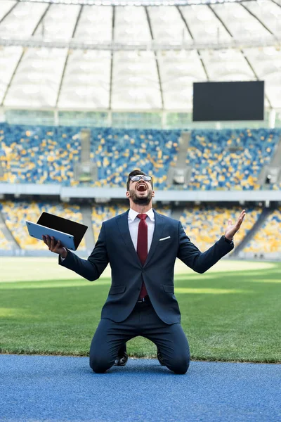 Happy young businessman in suit holding laptop with blank screen while standing on knees at stadium — Stock Photo