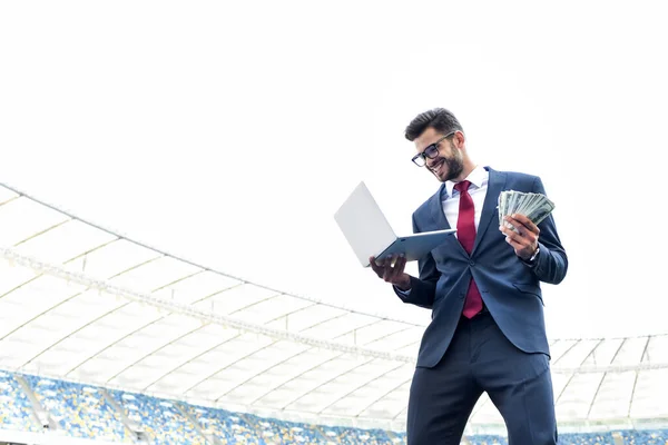 Low angle view of happy young businessman in suit holding laptop and money at stadium, sports betting concept — Stock Photo