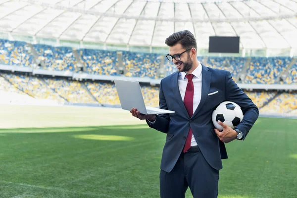 Sorridente jovem empresário de terno com laptop e bola de futebol no estádio, conceito de apostas esportivas — Fotografia de Stock