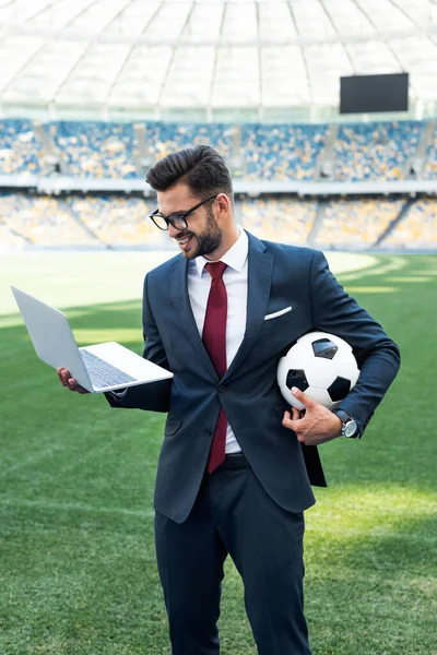 Улыбающийся молодой бизнесмен в костюме с ноутбуком и футбольным мячом на стадионе, концепция ставок на спорт — стоковое фото
