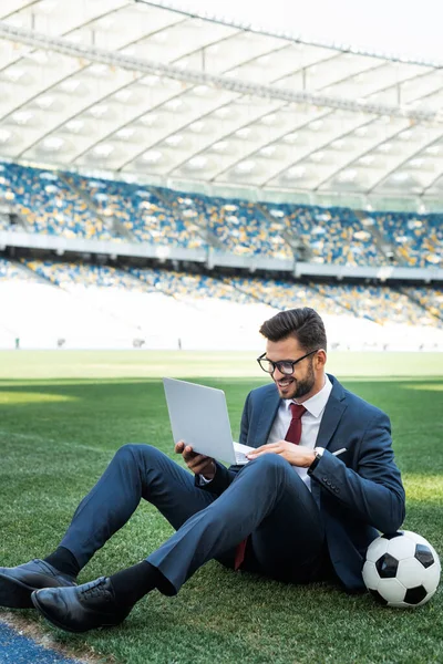 Sonriente joven hombre de negocios en traje con portátil y pelota de fútbol sentado en el campo de fútbol en el estadio, concepto de apuestas deportivas - foto de stock