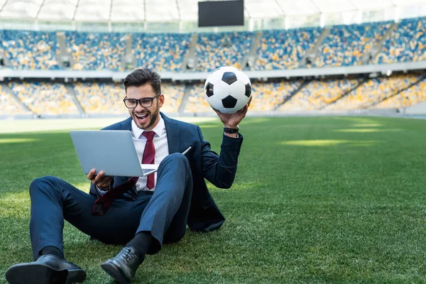 Feliz joven hombre de negocios en traje con portátil y pelota de fútbol sentado en el campo de fútbol en el estadio, concepto de apuestas deportivas - foto de stock