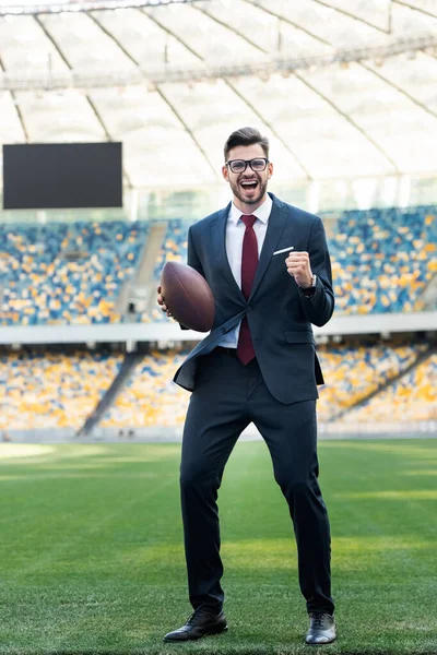 Счастливый молодой бизнесмен в костюме и очках с мячом для регби, показывающий жест 