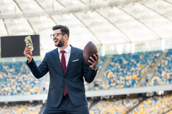 Счастливый молодой бизнесмен в костюме и очках с мячом для регби и деньгами на стадионе — стоковое фото