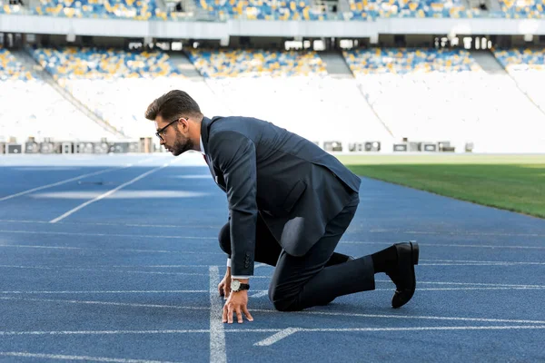 Вид сбоку молодого бизнесмена в костюме в стартовой позиции на беговой дорожке на стадионе — стоковое фото
