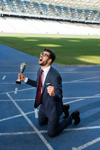 Jeune homme d'affaires en costume debout sur les genoux sur la piste de course avec trophée et crier au stade — Photo de stock